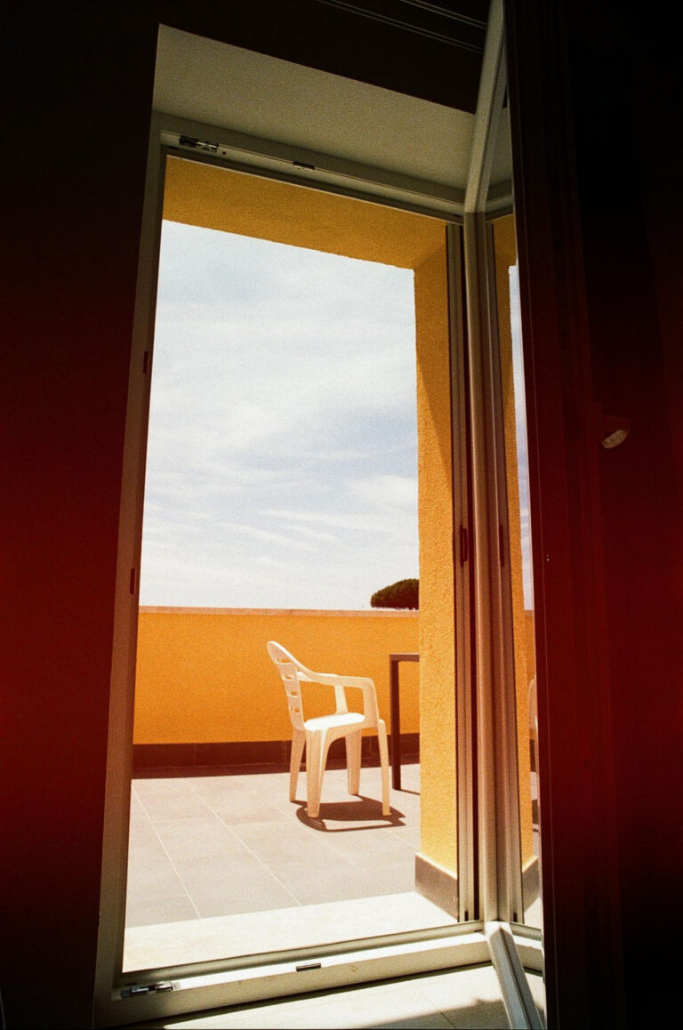 An open window framing a white chair on a sunlit balcony, the sea beyond