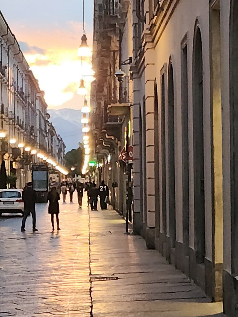 A porticoed street in Turin at sunset, hanging lamps lit, mountains on the horizon